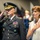 Lt. Gen. Dana Kyle Chipman retirement ceremony A U.S. Army general in dress uniform is rendering a salute during what appears to be a formal military ceremony. Beside him, a woman in a white lace dress has her right hand placed over her heart, suggesting the playing of the national anthem or a similar ceremonial moment. Color guard members in historical uniforms are visible in the background.