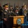 Lt. Gen. Dana Kyle Chipman retirement ceremony A senior U.S. Army general officer is delivering a speech at a formal military ceremony from a wooden podium bearing the Department of the Army seal. He is wearing a dress uniform with extensive medal ribbons, general officer rank insignia, and a service cap with gold braid. A military honor guard in ceremonial uniforms stands in formation behind him alongside multiple flags.