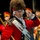 Lt. Gen. Dana Kyle Chipman retirement ceremony A man in 18th-century British Redcoat military costume is prominently featured in the foreground, appearing to perform in a historical reenactment or theatrical production. He wears a tall bearskin-style fur hat, red coat with gold epaulettes, white cross-belt, and carries a musket. Behind him are other performers in period and ceremonial military uniforms.