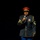 Lt. Gen. Dana Kyle Chipman retirement ceremony A man in a formal military dress uniform stands against a black background, speaking into a handheld microphone. He is wearing a dark jacket with gold trim, military medals, rank insignia, and a red-topped dress cap. The dramatic spotlight lighting isolates the subject, suggesting a formal performance or ceremonial event.
