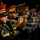Lt. Gen. Dana Kyle Chipman retirement ceremony A military band is performing, with multiple musicians in formal dress uniforms playing trumpets. The foreground features a sharp close-up of one musician in a black and gold uniform with a red-topped cap, actively playing his instrument. Additional band members are visible in the background in similar uniforms, slightly out of focus.