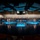 Lt. Gen. Dana Kyle Chipman retirement ceremony A large-scale military ceremonial event is taking place inside a grand indoor venue or auditorium. Hundreds of uniformed military personnel from what appear to be multiple branches are arranged in precise formations on a dark, reflective floor illuminated by blue spotlights. A row of flags lines the back of the stage, suggesting a formal joint-service or national ceremony.