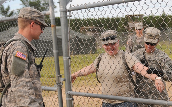 The image shows U.S. Army soldiers in ACU (Army Combat Uniform) interacting with a civilian at a chain-link fence gate, likely at a military installation or training area. One soldier on the left appears to be manning a checkpoint or entry control point, while a civilian in jeans and a tan shirt is being processed or escorted by another soldier. A fourth individual in uniform is visible in the background.
