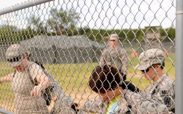 The image shows several U.S. Army soldiers in ACU (Army Combat Uniform) interacting with a child wearing a colorful shirt near a chain-link fence. The scene appears to take place on a military installation or training area, with open grassy fields and structures visible in the background. A civilian adult is also present on the left side of the frame.