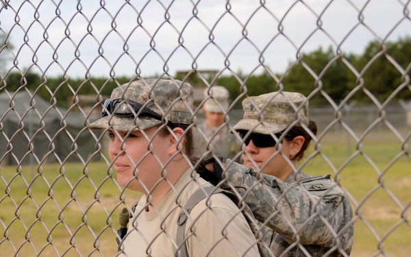 The image shows military personnel, appearing to be female soldiers, wearing U.S. Army ACU (Army Combat Uniform) pattern camouflage and carrying rucksacks, photographed through a chain-link fence. At least three individuals are visible, with two in the foreground and one in the background. The setting appears to be a military training installation or base.