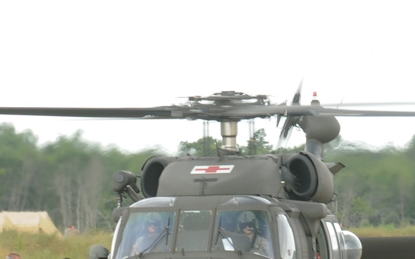 A military medical evacuation (MEDEVAC) helicopter, identified as a UH-60 Black Hawk with red cross markings, is on the ground in an open grassy field. Several military personnel in mixed uniforms are gathered near the aircraft, appearing to load or assist with a casualty or equipment. A field camp with tan tents is visible in the background.