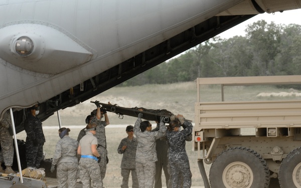 Military personnel in ABU (Airman Battle Uniform) camouflage are conducting cargo loading or unloading operations at the rear ramp of a US Air Force transport aircraft, identifiable by the USAF roundel insignia on the fuselage. A flatbed military truck trailer is positioned adjacent to the aircraft's open rear cargo ramp. The personnel appear to be manually handling a long piece of equipment or structural component between the aircraft and the truck.