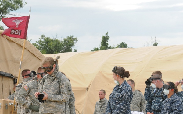 A joint military medical training exercise involving U.S. Army and U.S. Navy personnel is underway at a field medical station. Personnel in both Army ACU camouflage and Navy NWU uniforms are handling a patient on a wheeled litter/stretcher outside large tan military medical tents. The unit guidon flag marked '901' with a medical caduceus symbol identifies this as a medical unit exercise.