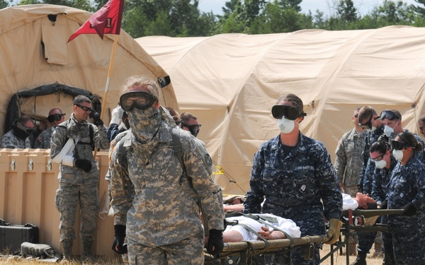 A military field exercise depicting a mass casualty or medical training drill, with personnel from multiple service branches (Army and Navy) transporting a simulated patient on a stretcher. Personnel are wearing protective goggles and face coverings, suggesting a CBRN (Chemical, Biological, Radiological, Nuclear) or pandemic response scenario. The setting includes multiple tan military field hospital tents with a red flag marker visible.