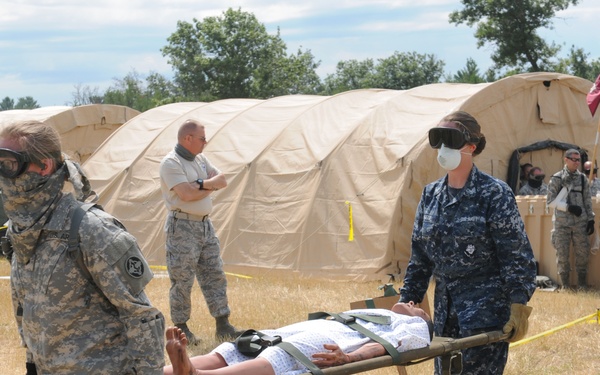 A military medical training exercise is underway at a field hospital setup with tan military tents. Two service members in different branch uniforms — Army ACU and Navy NWU — are carrying a simulated patient on a litter/stretcher. Multiple personnel in various uniforms and protective gear are visible in the background observing or participating in the exercise.