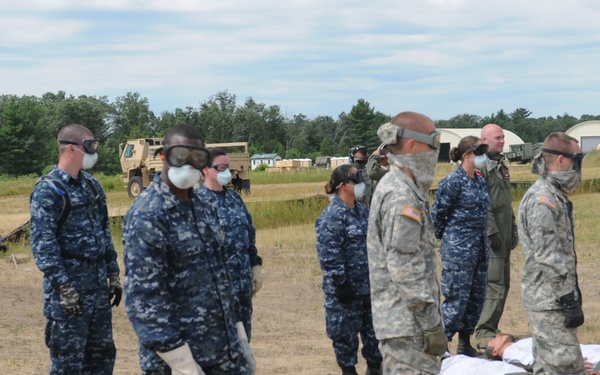 A joint military training exercise involving U.S. Army and U.S. Navy personnel conducting a mass casualty (MASCAL) drill in an outdoor field setting. Personnel are wearing protective gear including respirator masks and gloves while attending to simulated casualties on litters/stretchers. The scene depicts a CBRN (Chemical, Biological, Radiological, Nuclear) or medical triage training scenario.