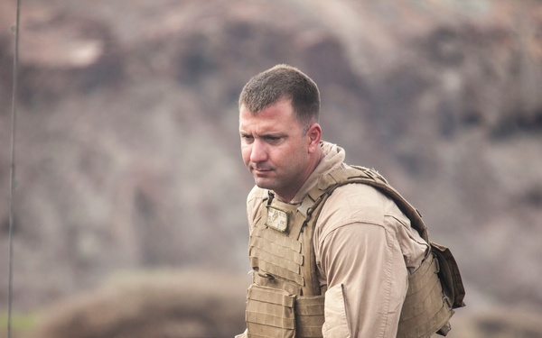 A male individual wearing tan/khaki military-style clothing and a tactical plate carrier vest is seated on what appears to be military equipment. A second individual in darker clothing with a vest is partially visible on the left side of the frame. The setting appears to be an outdoor, arid or rocky terrain environment.