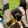 A person wearing black tactical/work gloves is holding a thin wire or cable with two components attached at each end — a small cylindrical blue glittery object at one end and a narrow metallic rod or tube at the other. The setting appears to be outdoors in a work or field environment, with green grass, a wooden post, and cardboard boxes visible in the background. The image is focused on the hand and the held components.