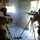 Two military personnel in desert camouflage uniforms are positioned inside a damaged structure, one operating a scoped rifle on a bipod and the other using an optical observation device on a tripod. A large debris cloud or blast effect is visible in the center of the image, consistent with a recent discharge or explosion through a wall. The setting appears to be a deteriorated building used as a firing or observation position.