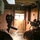 Two individuals are inside a dilapidated wooden structure, one holding a camera on a mount and the other holding what appears to be a rifle with an optic. A bright light source is visible through a small window in the interior door ahead of them. The scene appears to be a training or documentation exercise inside a derelict building.
