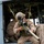 A military sniper in desert MARPAT camouflage uniform is positioned at the open door of a helicopter, aiming a scoped precision rifle outward. At least two other personnel are visible inside the aircraft, including what appears to be a flight crew member in a brown flight suit. The setting appears to be a grassy field, suggesting a training exercise or tactical insertion operation.