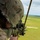 A military personnel wearing MARPAT camouflage and a combat helmet is firing a rifle from an elevated aerial position, likely from a helicopter doorway. A spent brass casing is visible ejecting from the weapon. The terrain below shows a flat, green open field consistent with a training range environment.