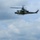 A U.S. Marine Corps UH-1Y Venom helicopter (tail number 32) is airborne over a military training range, with crew members visible in the open cabin doorway. Below on the ground, small target markers and at least one smoke plume are visible, indicating an active live-fire or aerial gunnery exercise. The setting appears to be a flat, grassy range bordered by a tree line under partly cloudy skies.