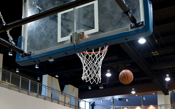 A wheelchair basketball game is in progress inside an indoor gymnasium. Player number 33 in a blue jersey is shooting the ball toward the basket, with the ball visible in mid-air near the hoop. Spectators fill the bleachers in the background, and a scoreboard shows the score as Guest 4, Home 6 with 10:58 remaining.