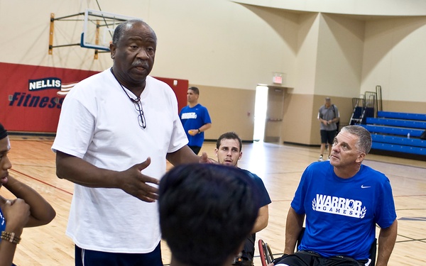 A coaching or instructional session is taking place at Nellis Fitness Center, where a standing man in a white t-shirt addresses a group of wheelchair basketball players. Several participants are wearing blue 'Warrior Games' athletic shirts, suggesting this is a military adaptive sports program. The group is gathered in a circle on an indoor basketball court, attentively listening to the instructor.
