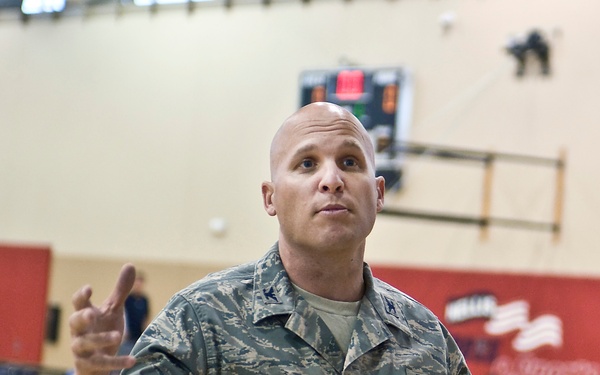 A U.S. Air Force servicemember named Perham, wearing ABU (Airman Battle Uniform) camouflage, is speaking or addressing someone in a gymnasium setting. In the background, individuals in wheelchairs are visible on a basketball court, suggesting a wheelchair basketball event or adaptive sports activity. The subject appears to be gesturing expressively with both hands raised.