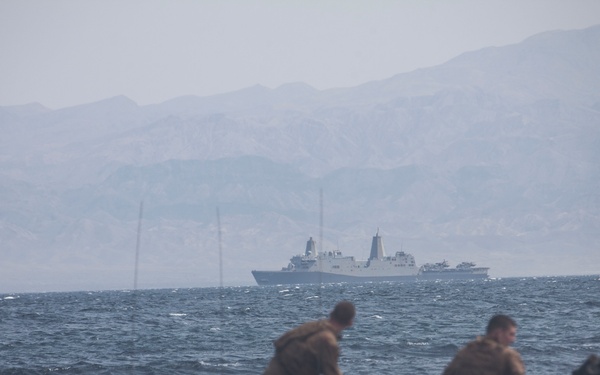 The image shows military personnel aboard amphibious assault vehicles (AAVs) operating in open water, likely conducting an amphibious landing exercise. A large naval vessel, consistent with a San Antonio-class amphibious transport dock ship, is visible in the background. Mountainous terrain is visible through haze on the horizon.