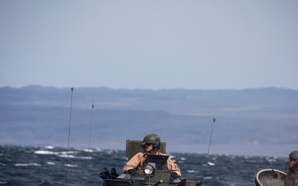 An amphibious assault vehicle (AAV) is navigating through choppy open water with at least three military personnel visible on board. The vehicle appears to be a US Marine Corps AAV7A1, identifiable by its distinctive hull shape and olive drab coloring. A coastline with hills is visible in the background, suggesting a maritime training exercise or amphibious landing operation.