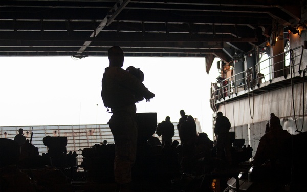 A silhouetted scene inside what appears to be a naval vessel's well deck or cargo bay, with multiple personnel visible against a bright exterior opening. The image shows military personnel carrying gear and equipment in a darkened interior space alongside what appear to be armored or military vehicles. The strong backlight from the open bay door creates dramatic silhouettes throughout the scene.