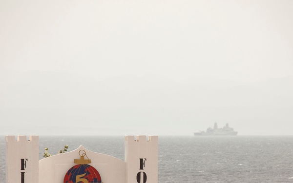 The image shows a military installation entrance gate with the text 'FIER FORT' and a unit emblem featuring the number 5, an anchor, and a globe design. An armed soldier in camouflage uniform stands guard to the left of the white castle-style gate structure. In the background, a naval vessel is visible on the open sea under hazy atmospheric conditions.