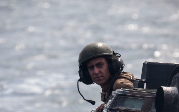 A military personnel member is visible from the hatch of an amphibious armored vehicle traversing open water. The soldier is wearing a combat helmet and communications headset, suggesting active operational or training activity. The vehicle appears to be an amphibious assault vehicle (AAV) or similar tracked amphibious platform.