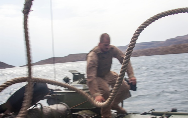 The image shows military personnel on top of what appears to be an amphibious assault vehicle (AAV) operating on open water. A service member in tan/desert camouflage is handling a large rope or hawser line on the vehicle's hull. The scene appears to depict a maritime training or operational exercise, with hilly terrain visible in the background.