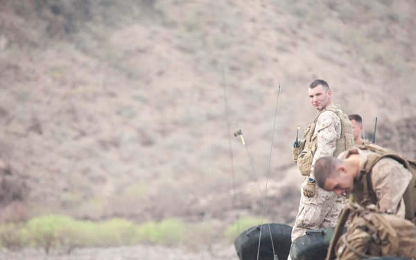 A group of military personnel in desert camouflage uniforms are gathered around military vehicles loaded with tactical gear and equipment. The soldiers appear to be staging, loading, or preparing equipment in an arid, hilly terrain environment. The scene suggests a field operation, training exercise, or mission preparation.