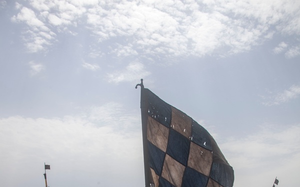 Military personnel aboard a watercraft are conducting a maritime operation, with one individual holding a large blue and tan checkered flag. Multiple vessels are visible in the water near a rocky coastline, suggesting a coordinated naval exercise or amphibious operation. The scene appears to be a training or tactical maneuver in a coastal environment.