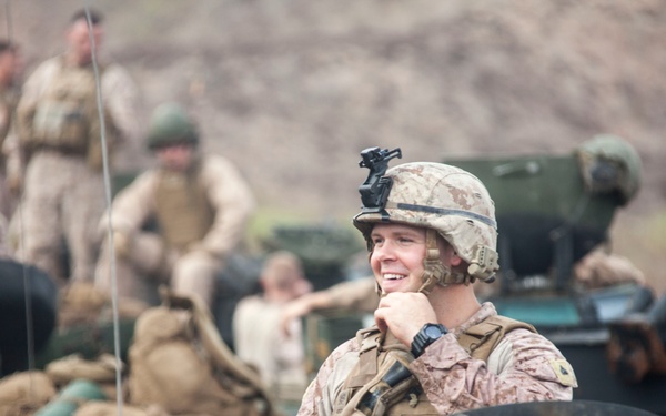 A smiling military service member in MARPAT (Marine pattern) camouflage uniform and helmet is positioned in or near a military vehicle, surrounded by gear and equipment. Several other service members are visible in the background in a field or training environment. The scene appears to depict a military exercise or operational staging area.