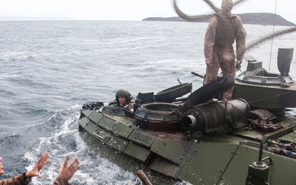 Military personnel are operating amphibious assault vehicles (AAVs) on open ocean water. At least three soldiers in combat uniforms are visible across two vehicles, with one soldier appearing to gesture or reach toward the adjacent vehicle. The scene depicts a maritime military operation or training exercise, with a distant landmass visible on the horizon.