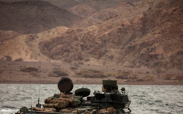 An amphibious assault vehicle (AAV) is traversing open water, likely during a military exercise or amphibious landing operation. The vehicle is heavily laden with equipment and gear on its top deck, moving through choppy water with visible wake and spray. Arid, rocky mountains are visible in the background, suggesting a desert coastal environment.