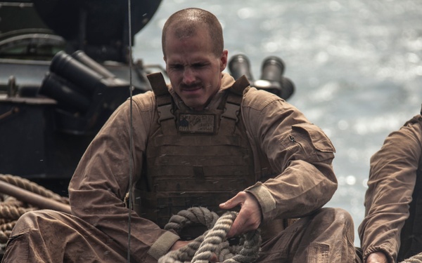 Two military personnel in tan/desert camouflage uniforms and tactical vests are handling thick rope lines aboard a naval vessel on open water. The central figure is actively managing a coiled rope while seated on the deck of what appears to be a landing craft or patrol boat. The scene suggests a maritime mooring, docking, or vessel-to-vessel transfer operation.