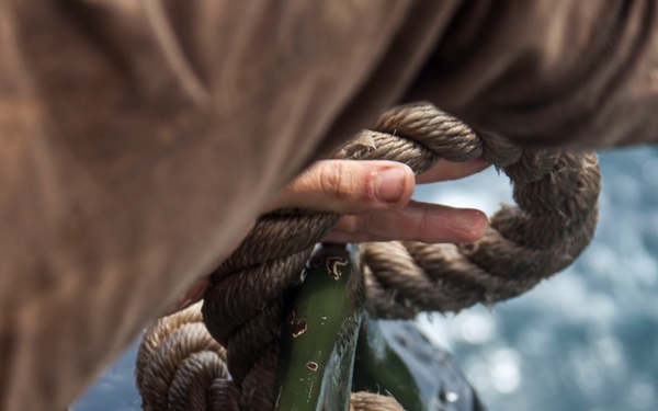 A close-up photograph of a person's hand working with a thick, weathered hemp or natural fiber rope wrapped around a green-painted metal cleat on what appears to be a boat or vessel. The rope shows significant wear and moisture, suggesting active maritime use. Water is visible in the background, confirming a marine environment.
