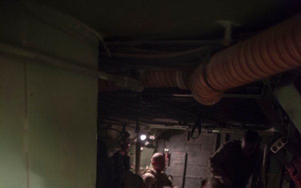Military personnel in desert camouflage uniforms are seated inside what appears to be the cargo hold or interior of a military aircraft or large vehicle. The space is dimly lit and cramped, with visible ducting, equipment panels, and gear. Personnel appear to be in transit or staging for an operation.