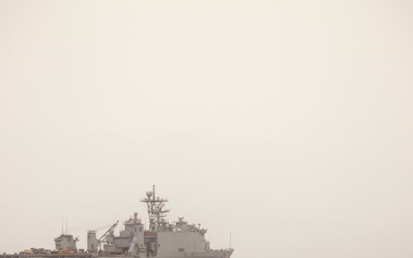 The image shows a military amphibious assault vehicle (AAV) operating in open ocean waters, with a large naval warship visible in the background. The scene appears to depict an amphibious military exercise or operation, with the AAV moving through choppy seas away from or toward the ship. Hazy atmospheric conditions reduce overall visibility.