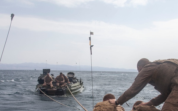 Military personnel in tan/coyote brown uniforms are conducting a maritime operation involving two amphibious assault vehicles (AAVs) connected by a rope or tow line on open water. Soldiers on the near vessel are actively handling the line connecting to the second vehicle in the distance. The scene appears to be a training or operational exercise with choppy sea conditions and mountainous coastline visible in the background.