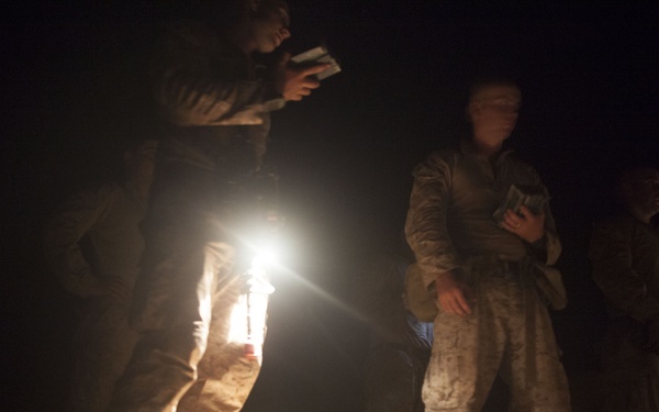 A nighttime scene showing military personnel in desert camouflage uniforms gathered around a light source on the ground. The soldiers appear to be reviewing documents or maps by the light of what appears to be a lantern or flashlight. Military gear and equipment bags are visible on the ground between them.