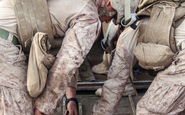 The image shows multiple military personnel in desert camouflage uniforms bending over and working on mechanical equipment, likely securing or connecting a chain to a coupling or towing mechanism. The soldiers appear to be performing a maintenance or rigging task on what looks like a military vehicle or equipment platform. The photo is taken from a top-down perspective focusing on the lower torsos and hands of the personnel.