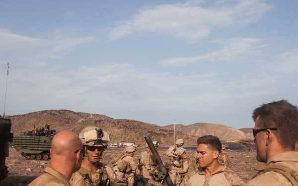 A group of military personnel in desert camouflage uniforms are gathered in a rocky, arid coastal environment, appearing to be in discussion or receiving a briefing. Behind them, an artillery piece and armored amphibious assault vehicles are visible. The setting suggests a military exercise or operational deployment near a coastline.