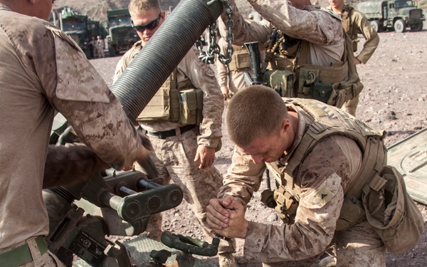 A group of military personnel in desert camouflage uniforms are working together to assemble or position what appears to be an artillery piece or towed weapon system. The scene takes place in an arid, rocky terrain with military vehicles visible in the background. The soldiers appear to be engaged in a field training exercise or operational deployment.