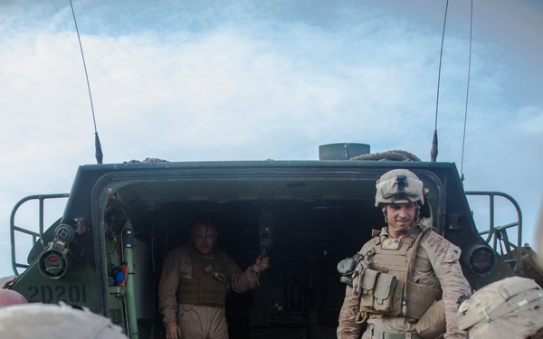 Military personnel in desert camouflage (MARPAT) uniforms are operating on what appears to be a Landing Craft Air Cushion (LCAC) or similar military amphibious vessel. Several Marines are visible on the deck alongside what appears to be an artillery piece or towed weapon system. One individual in tan/khaki uniform stands in the vehicle bay opening while others in full combat gear move about the deck.