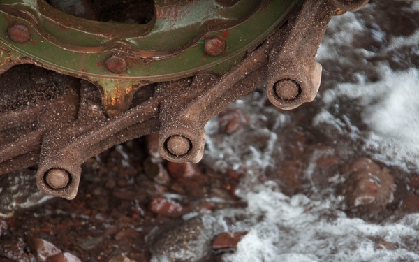 The image shows heavily corroded and rusted mechanical machinery, likely part of a shipwreck or industrial equipment, resting on a rocky shoreline. Ocean waves with white foam are washing over and around the metal components and pebbled beach. The machinery appears to have been exposed to saltwater for an extended period, evidenced by severe oxidation and encrustation.