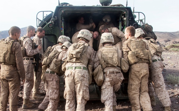 A group of military personnel in desert camouflage uniforms are gathered around the rear of a military tactical vehicle, with several soldiers appearing to load or unload something through the open rear hatch. The setting is an arid, rocky desert terrain with sparse vegetation and hills visible in the background. The soldiers are wearing full combat gear including helmets, body armor, and tactical vests.