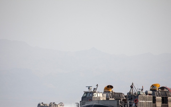 Two U.S. Navy Landing Craft Air Cushion (LCAC) hovercraft are operating at speed on open water with mountainous terrain visible in the background. The foreground LCAC bears the number '02' and displays a U.S. flag, along with yellow and checkered markings typical of naval hovercraft. The craft are generating spray and appear to be conducting a transit or amphibious exercise.