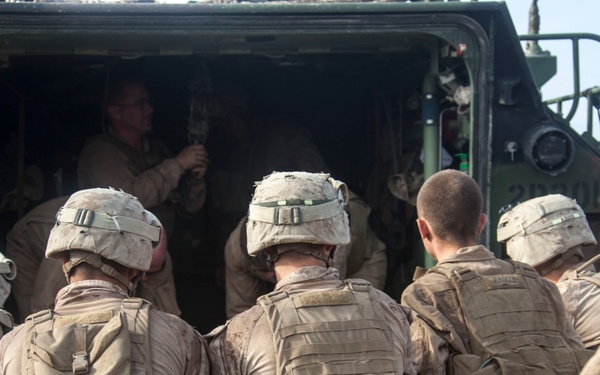 A group of military personnel in full combat gear are boarding or loading into a large military vehicle. The soldiers are wearing desert camouflage uniforms, tactical vests, and helmets, suggesting a training exercise or operational deployment. The scene appears to take place in an arid or desert environment.